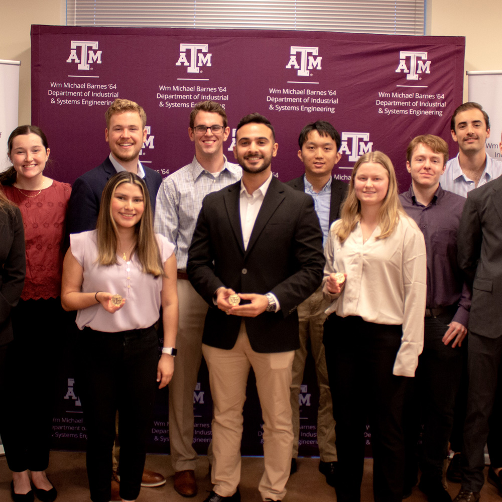 A group of students stand in front of a maroon background. All hold small gold coins.