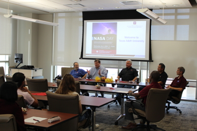 五个men sit at a table, talking with a room of students and faculty. A NASA Day informational slide is on a screen behind the men.